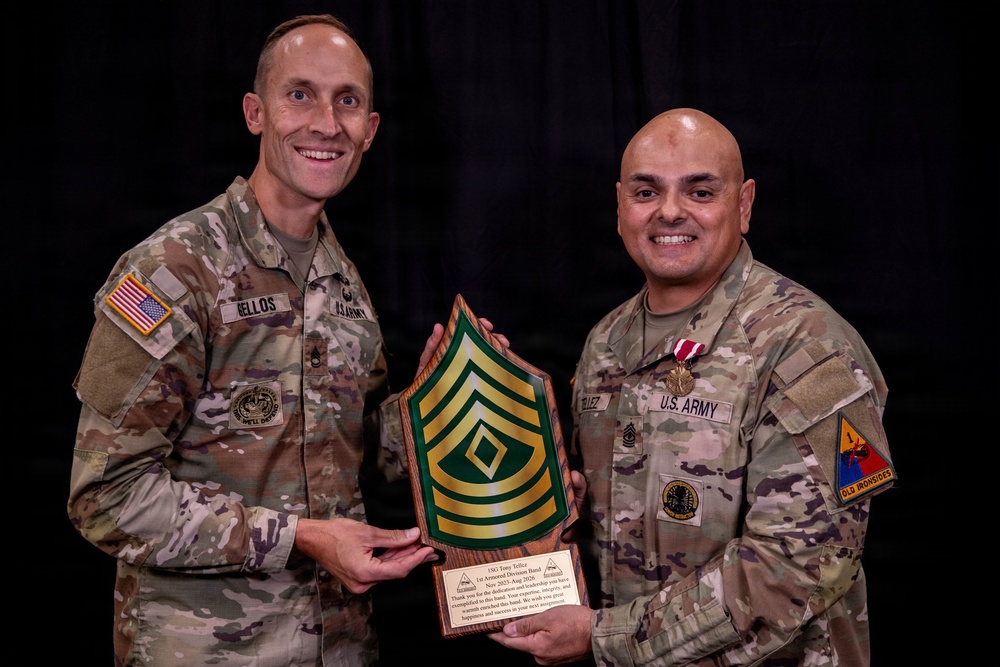Two U.S. Army soldiers in OCP (Operational Camouflage Pattern) uniforms pose together holding a wooden plaque featuring a First Sergeant rank insignia (chevrons with a diamond). The soldier on the right, identified by nametape as 'TELLEZ,' is wearing a medal and a '1st Armored Division' (Old Ironsides) shoulder sleeve insignia. This appears to be a military recognition or farewell ceremony.
