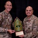 Two U.S. Army soldiers in OCP (Operational Camouflage Pattern) uniforms pose together holding a wooden plaque featuring a First Sergeant rank insignia (chevrons with a diamond). The soldier on the right, identified by nametape as 'TELLEZ,' is wearing a medal and a '1st Armored Division' (Old Ironsides) shoulder sleeve insignia. This appears to be a military recognition or farewell ceremony.