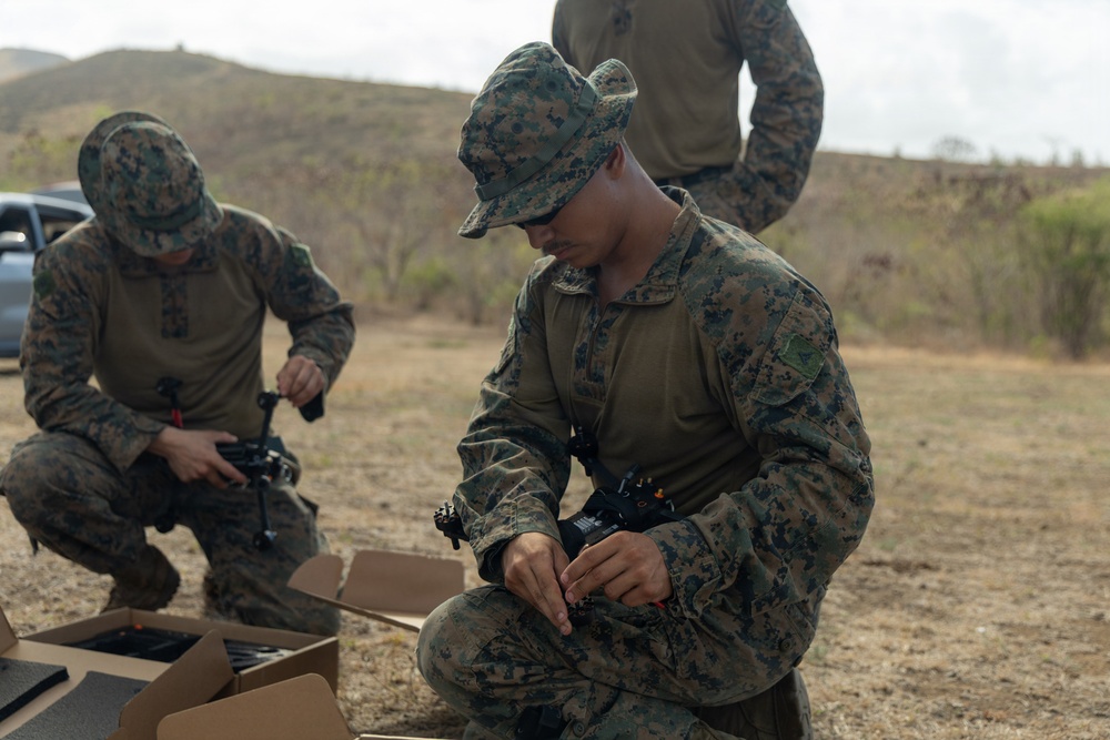 Two U.S. Marines in MARPAT digital camouflage uniforms are kneeling on dry ground assembling or preparing small drone/UAS (Unmanned Aerial System) equipment. A third Marine is partially visible standing in the background. The setting appears to be an outdoor training area with arid terrain and hills in the background.