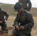 Two U.S. Marines in MARPAT digital camouflage uniforms are kneeling on dry ground assembling or preparing small drone/UAS (Unmanned Aerial System) equipment. A third Marine is partially visible standing in the background. The setting appears to be an outdoor training area with arid terrain and hills in the background.