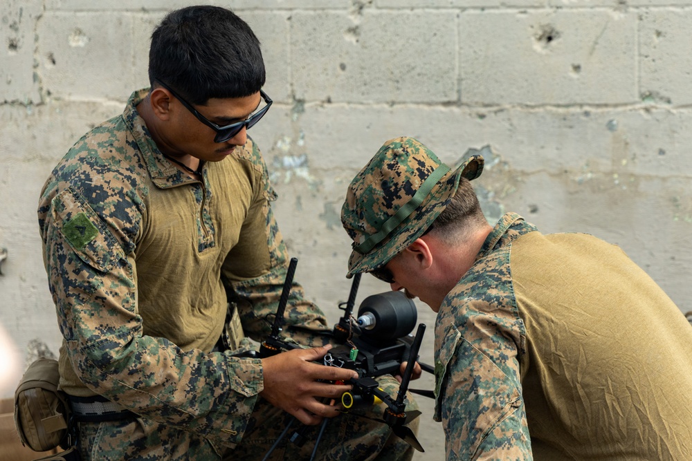Two military personnel in digital camouflage uniforms are working together to inspect or assemble a small drone. One individual is standing and holding the drone while the other leans in closely to examine a component. The setting appears to be near a concrete block wall, suggesting a field or training environment.