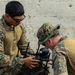 Two military personnel in digital camouflage uniforms are working together to inspect or assemble a small drone. One individual is standing and holding the drone while the other leans in closely to examine a component. The setting appears to be near a concrete block wall, suggesting a field or training environment.