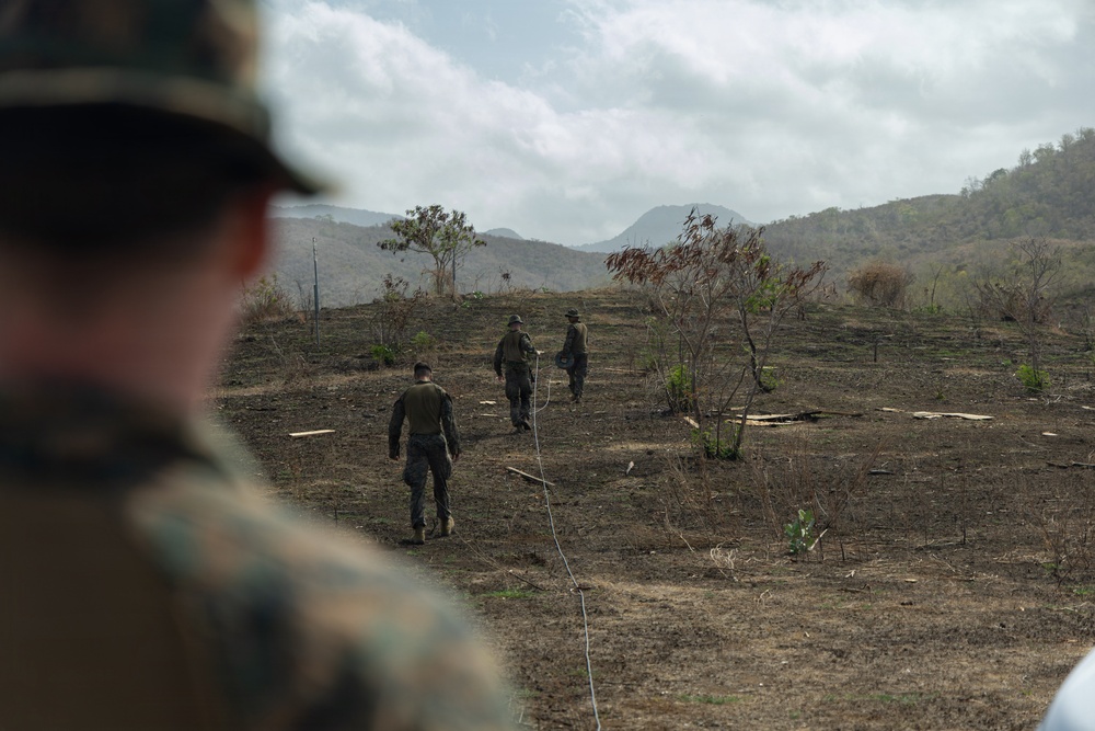 The image shows military personnel in camouflage uniforms walking across a cleared, partially burned hillside terrain. Three soldiers are visible moving away from the camera into the field, while a fourth soldier in the foreground is observed from behind. The setting appears to be a hilly, semi-arid landscape with sparse vegetation and mountains in the background.