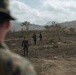 The image shows military personnel in camouflage uniforms walking across a cleared, partially burned hillside terrain. Three soldiers are visible moving away from the camera into the field, while a fourth soldier in the foreground is observed from behind. The setting appears to be a hilly, semi-arid landscape with sparse vegetation and mountains in the background.