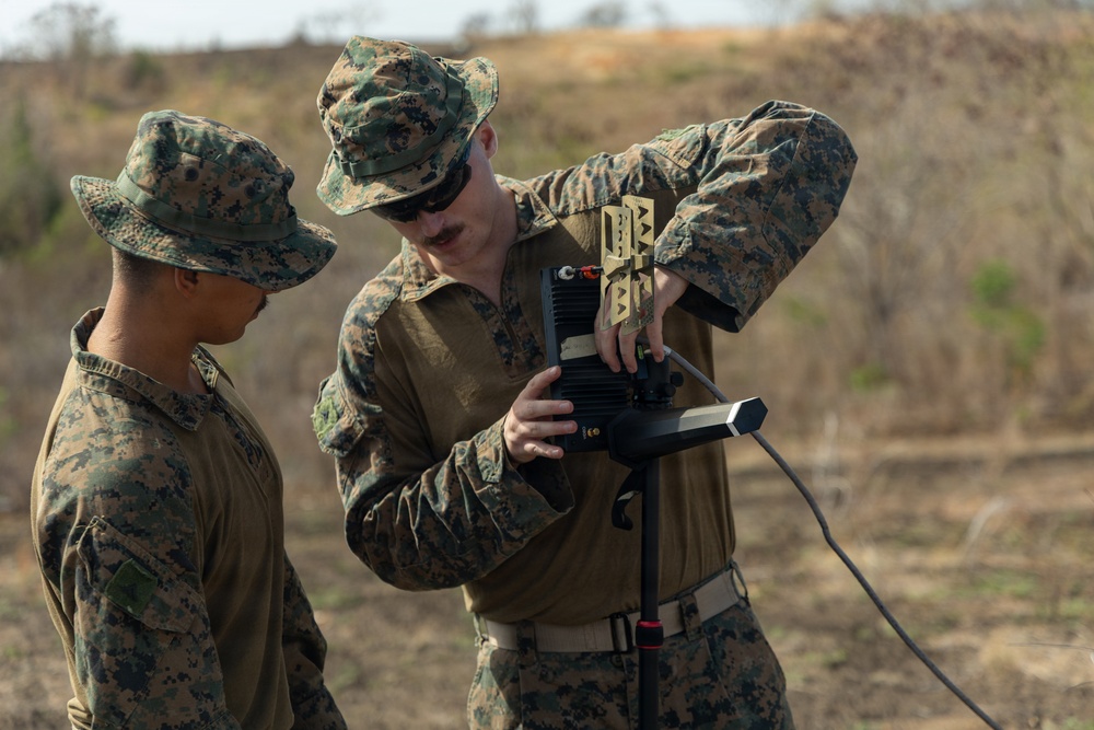 Two military personnel in MARPAT (Marine pattern) digital camouflage uniforms are working with a piece of electronic or communications equipment mounted on a tripod in an outdoor field environment. One individual appears to be instructing or demonstrating the operation of the device to the other. The setting appears to be a training exercise in an arid or semi-arid terrain.