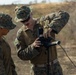 Two military personnel in MARPAT (Marine pattern) digital camouflage uniforms are working with a piece of electronic or communications equipment mounted on a tripod in an outdoor field environment. One individual appears to be instructing or demonstrating the operation of the device to the other. The setting appears to be a training exercise in an arid or semi-arid terrain.