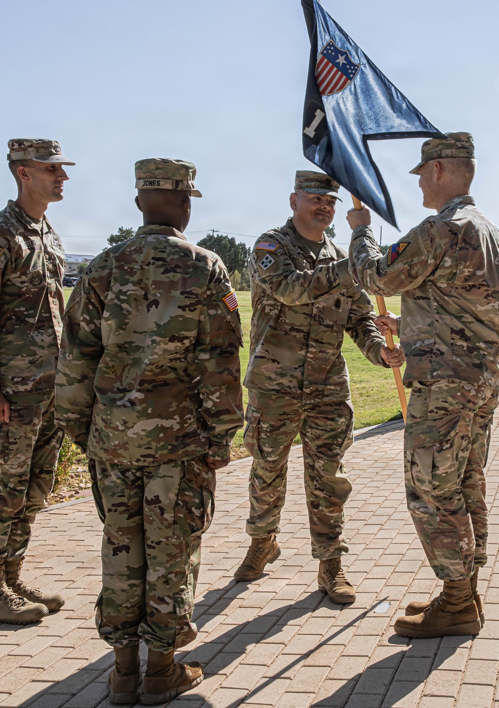 A U.S. Army military ceremony is taking place outdoors on a brick walkway, likely a change of command or activation ceremony. Four soldiers in OCP (Operational Camouflage Pattern) uniforms are participating, with two soldiers in the center exchanging or passing a unit guidon (flag). The guidon is black and features a shield with stars and stripes, along with the number '1', suggesting a first unit designation.