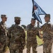 A U.S. Army military ceremony is taking place outdoors on a brick walkway, likely a change of command or activation ceremony. Four soldiers in OCP (Operational Camouflage Pattern) uniforms are participating, with two soldiers in the center exchanging or passing a unit guidon (flag). The guidon is black and features a shield with stars and stripes, along with the number '1', suggesting a first unit designation.