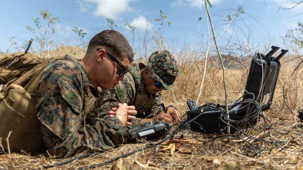 Two military personnel in MARPAT camouflage uniforms are lying prone in a dry, grassy field while operating communications or electronic equipment. They appear to be conducting a field exercise involving a portable electronic device and a hard-shell equipment case with cables. The setting suggests a training environment in an arid or semi-arid outdoor location.