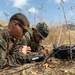Two military personnel in MARPAT camouflage uniforms are lying prone in a dry, grassy field while operating communications or electronic equipment. They appear to be conducting a field exercise involving a portable electronic device and a hard-shell equipment case with cables. The setting suggests a training environment in an arid or semi-arid outdoor location.