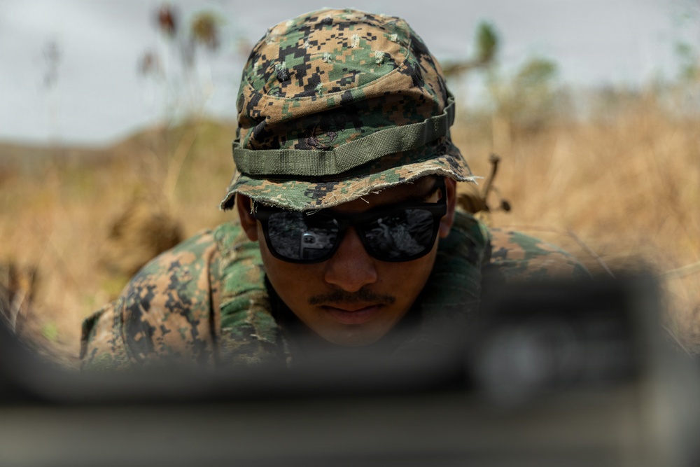 A military service member wearing digital camouflage (MARPAT pattern) utilities and a boonie cover is photographed in a low prone or crouched position in an outdoor field environment. The individual is wearing dark tactical sunglasses and appears to be observing or aiming over a blurred foreground object, likely a weapon or barrier. The background shows dry grassland vegetation under an overcast sky.
