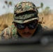 A military service member wearing digital camouflage (MARPAT pattern) utilities and a boonie cover is photographed in a low prone or crouched position in an outdoor field environment. The individual is wearing dark tactical sunglasses and appears to be observing or aiming over a blurred foreground object, likely a weapon or barrier. The background shows dry grassland vegetation under an overcast sky.