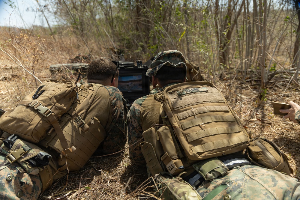Two military personnel in MARPAT (Marine pattern) camouflage uniforms and tan tactical vests are lying prone in a dry, brushy outdoor environment. They appear to be observing or operating a tactical display screen or equipment positioned in front of them. The 'MARINE' tape is visible on one of the vests, identifying them as U.S. Marines.