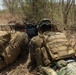 Two military personnel in MARPAT (Marine pattern) camouflage uniforms and tan tactical vests are lying prone in a dry, brushy outdoor environment. They appear to be observing or operating a tactical display screen or equipment positioned in front of them. The 'MARINE' tape is visible on one of the vests, identifying them as U.S. Marines.