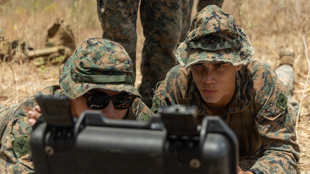 Two military personnel in MARPAT woodland camouflage uniforms and boonie covers are prone on dry ground, closely examining a piece of military equipment in the foreground. A third individual is partially visible standing behind them. The setting appears to be an outdoor training environment with dry, arid vegetation.