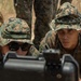 Two military personnel in MARPAT woodland camouflage uniforms and boonie covers are prone on dry ground, closely examining a piece of military equipment in the foreground. A third individual is partially visible standing behind them. The setting appears to be an outdoor training environment with dry, arid vegetation.