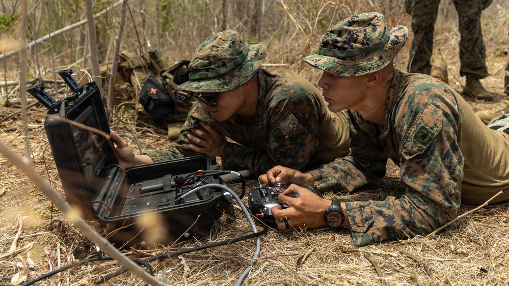 Two military personnel in MARPAT woodland camouflage uniforms are lying prone on dry ground, operating what appears to be a drone ground control station with a monitor and handheld controller. They are in a sparse, dry woodland environment, intently focused on a screen displaying what is likely a live drone feed. A third individual is partially visible standing in the background.