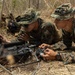 Two military personnel in MARPAT woodland camouflage uniforms are lying prone on dry ground, operating what appears to be a drone ground control station with a monitor and handheld controller. They are in a sparse, dry woodland environment, intently focused on a screen displaying what is likely a live drone feed. A third individual is partially visible standing in the background.