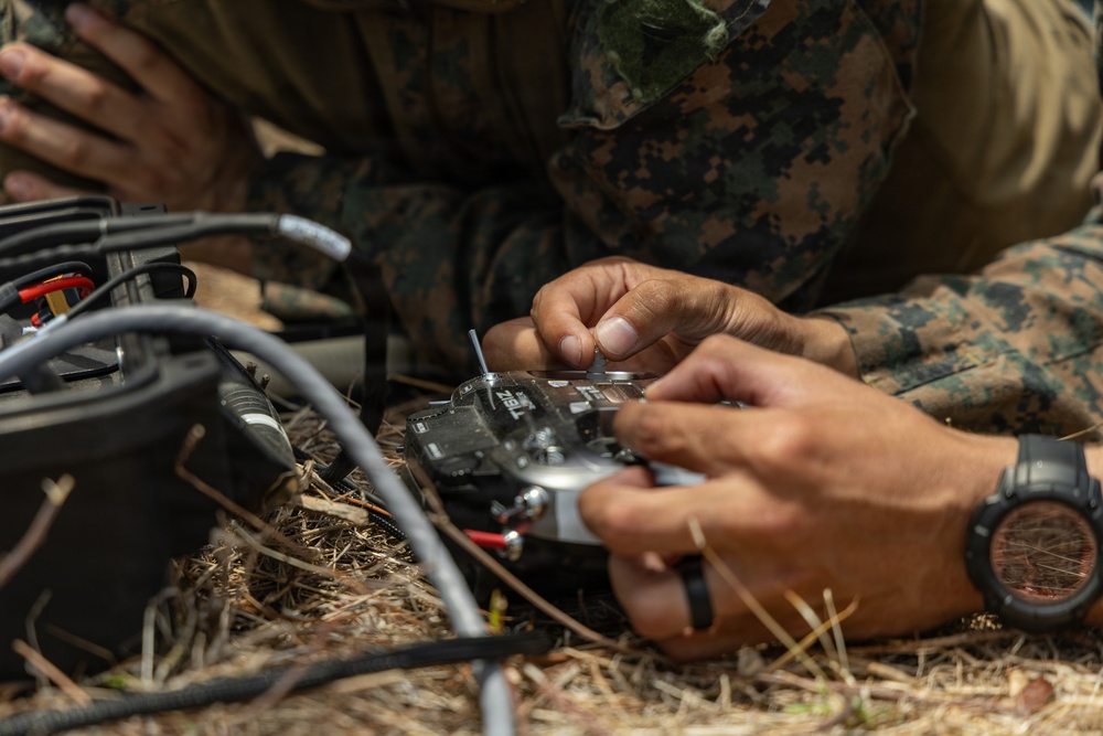 Military personnel in camouflage uniforms are working with electronic equipment on the ground in an outdoor, wooded setting. They appear to be handling a radio transmitter or drone remote control unit along with associated cables and wiring. The close-up perspective emphasizes the hands-on technical work being performed.