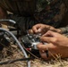 Military personnel in camouflage uniforms are working with electronic equipment on the ground in an outdoor, wooded setting. They appear to be handling a radio transmitter or drone remote control unit along with associated cables and wiring. The close-up perspective emphasizes the hands-on technical work being performed.
