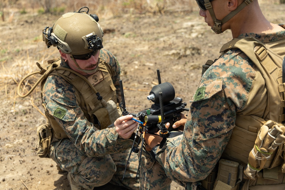 Two military personnel in MARPAT (Marine pattern) camouflage uniforms are handling a small drone or unmanned aerial system (UAS) in an outdoor field environment. The soldier on the left is wearing a tactical helmet with mounted devices and is actively working on or inspecting the drone. The setting appears to be a dry, arid training or operational area.