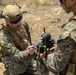 Two military personnel in MARPAT (Marine pattern) camouflage uniforms are handling a small drone or unmanned aerial system (UAS) in an outdoor field environment. The soldier on the left is wearing a tactical helmet with mounted devices and is actively working on or inspecting the drone. The setting appears to be a dry, arid training or operational area.