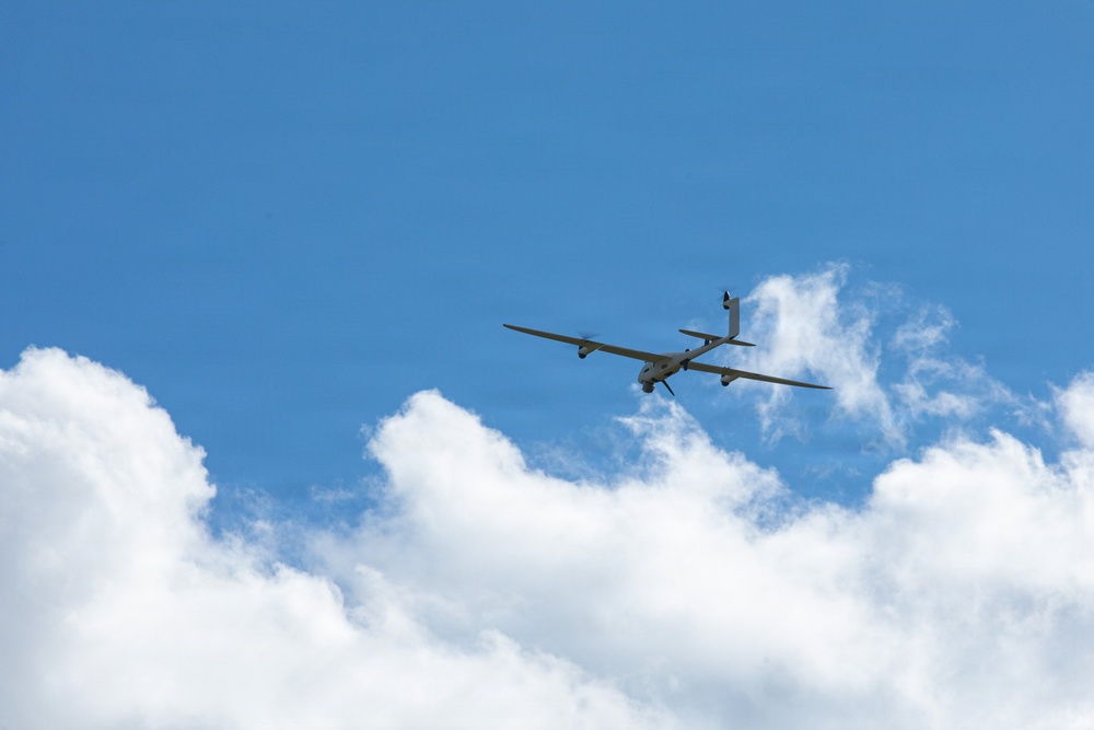 An unmanned aerial vehicle (UAV) or drone is captured in flight against a bright blue sky with scattered white cumulus clouds. The aircraft has a distinctive long-wingspan glider-like design with a pusher propeller configuration, consistent with a military or surveillance-type UAV. The image is taken from a low angle looking upward, providing a clear silhouette of the aircraft.