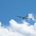 An unmanned aerial vehicle (UAV) or drone is captured in flight against a bright blue sky with scattered white cumulus clouds. The aircraft has a distinctive long-wingspan glider-like design with a pusher propeller configuration, consistent with a military or surveillance-type UAV. The image is taken from a low angle looking upward, providing a clear silhouette of the aircraft.