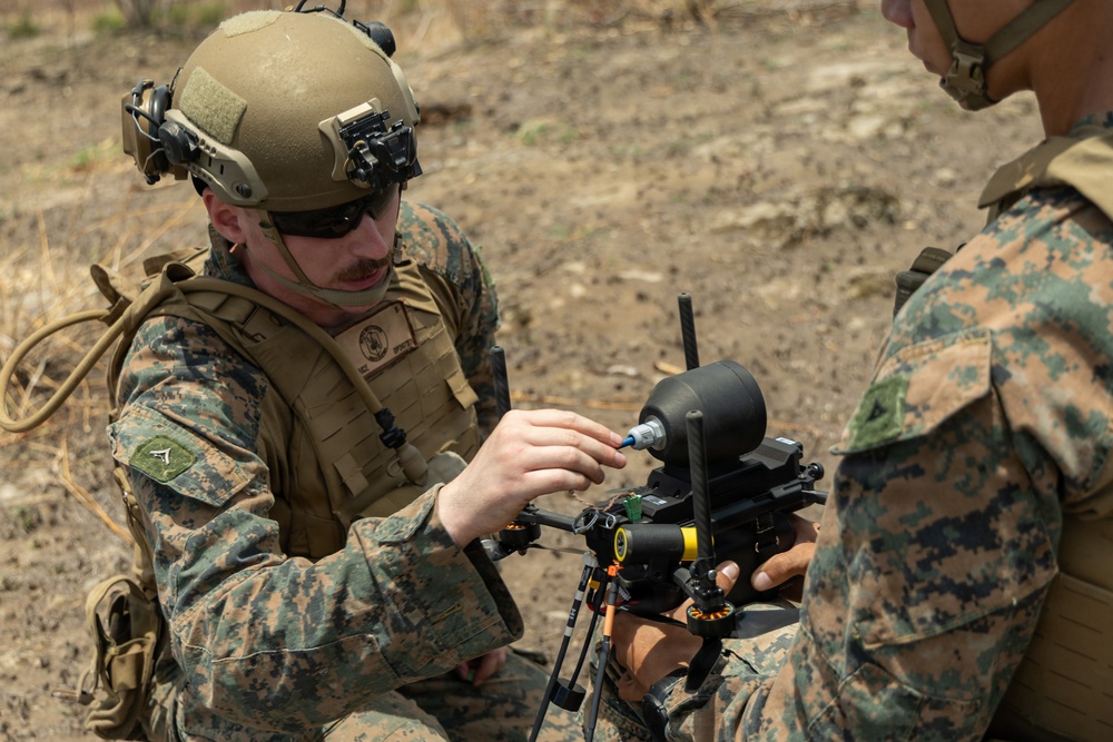 Two military personnel in MARPAT (Marine pattern) camouflage uniforms are working together to assemble or configure a small drone/UAS (Unmanned Aerial System) in an outdoor field environment. The soldier in the foreground is wearing a tactical helmet with night-vision mount and is connecting what appears to be a cable or component to the drone. The setting appears to be a dry, arid training or operational area.