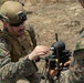 Two military personnel in MARPAT (Marine pattern) camouflage uniforms are working together to assemble or configure a small drone/UAS (Unmanned Aerial System) in an outdoor field environment. The soldier in the foreground is wearing a tactical helmet with night-vision mount and is connecting what appears to be a cable or component to the drone. The setting appears to be a dry, arid training or operational area.