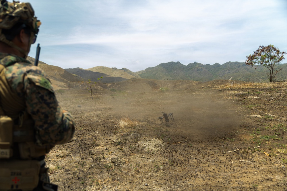 A military service member in MARPAT camouflage uniform observes a field exercise from the foreground, while a dust cloud rises around what appears to be a mortar or weapon system firing in the middle distance. The terrain is arid and open, with rolling hills and mountains visible in the background. The scene is consistent with a live-fire training exercise conducted in a dry, tropical or subtropical environment.
