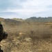 A military service member in MARPAT camouflage uniform observes a field exercise from the foreground, while a dust cloud rises around what appears to be a mortar or weapon system firing in the middle distance. The terrain is arid and open, with rolling hills and mountains visible in the background. The scene is consistent with a live-fire training exercise conducted in a dry, tropical or subtropical environment.