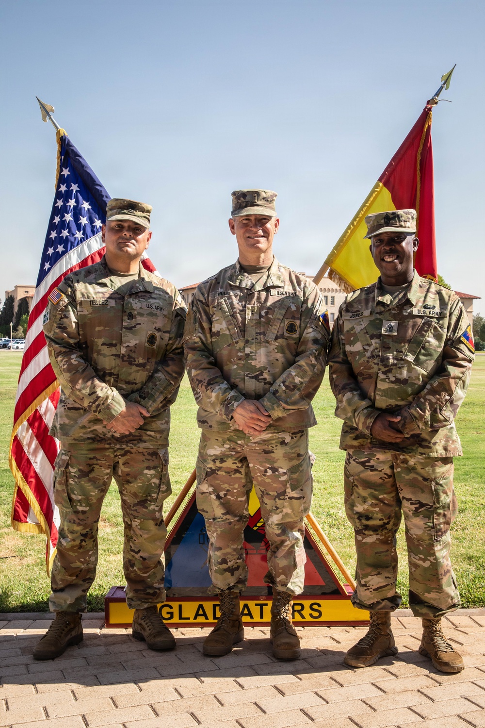 Three U.S. Army soldiers in OCP (Operational Camouflage Pattern) uniforms pose for a formal photograph in front of an American flag and a military unit guidon. Name tapes visible on the uniforms read 'TELLEZ' (left), 'HULL' (center), and 'JONES' (right). They are standing in front of a sign reading 'GLADIATORS' on what appears to be a military installation parade field.