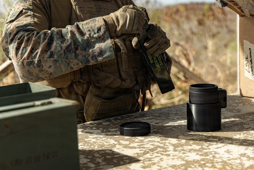 A military personnel wearing MARPAT (Marine pattern) camouflage and tactical gear is standing at an outdoor table, handling what appears to be a black cylindrical container with its lid removed. The setting appears to be a training range or field environment, with an ammunition can visible on the left side of the table. The individual is wearing tactical gloves and a plate carrier vest with pouches.
