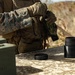 A military personnel wearing MARPAT (Marine pattern) camouflage and tactical gear is standing at an outdoor table, handling what appears to be a black cylindrical container with its lid removed. The setting appears to be a training range or field environment, with an ammunition can visible on the left side of the table. The individual is wearing tactical gloves and a plate carrier vest with pouches.