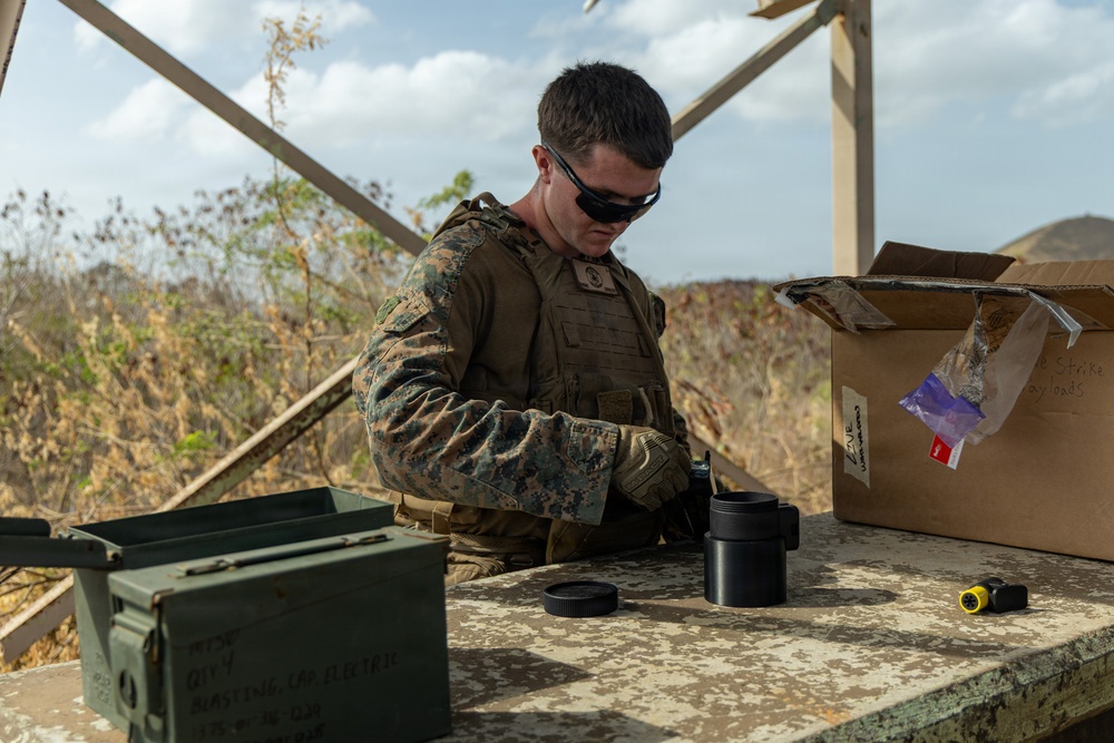 A U.S. Marine in MARPAT camouflage uniform and tactical vest is working at an outdoor table, handling what appears to be optical or cylindrical equipment components. The setting appears to be a forward operating or training area with dry, arid vegetation visible in the background. A cardboard box partially labeled 'Strike Payloads' and a military ammunition can are visible on the table.