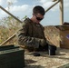 A U.S. Marine in MARPAT camouflage uniform and tactical vest is working at an outdoor table, handling what appears to be optical or cylindrical equipment components. The setting appears to be a forward operating or training area with dry, arid vegetation visible in the background. A cardboard box partially labeled 'Strike Payloads' and a military ammunition can are visible on the table.