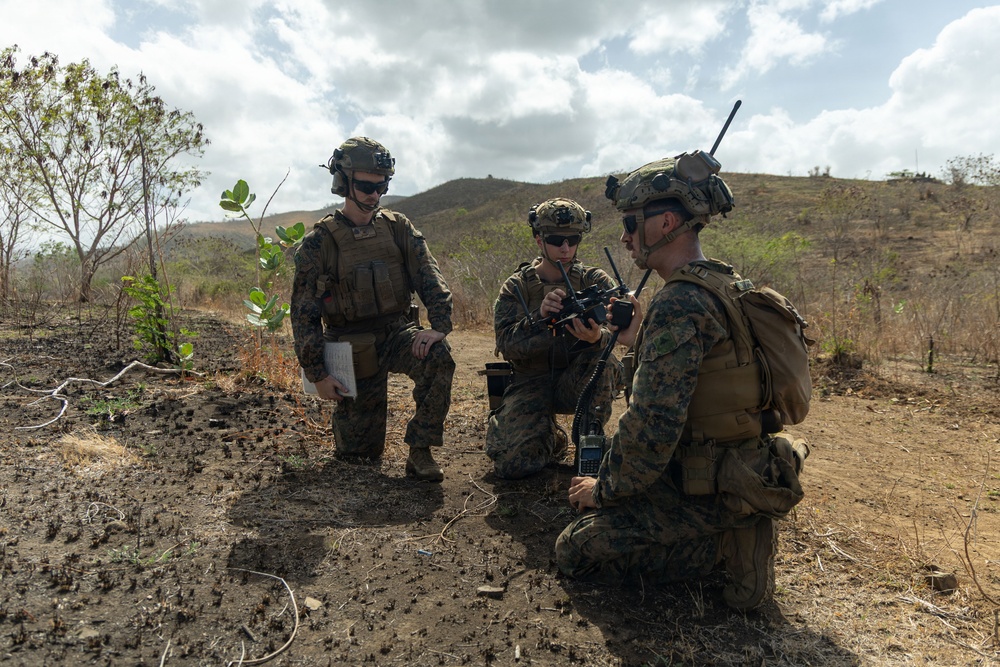 Three military personnel in camouflage uniforms and tactical gear are gathered in an arid, hilly outdoor environment. Two are kneeling while operating radio/communication equipment, and one is standing holding what appears to be a notepad or map. The setting suggests a field training exercise or operational mission.