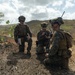 Three military personnel in camouflage uniforms and tactical gear are gathered in an arid, hilly outdoor environment. Two are kneeling while operating radio/communication equipment, and one is standing holding what appears to be a notepad or map. The setting suggests a field training exercise or operational mission.
