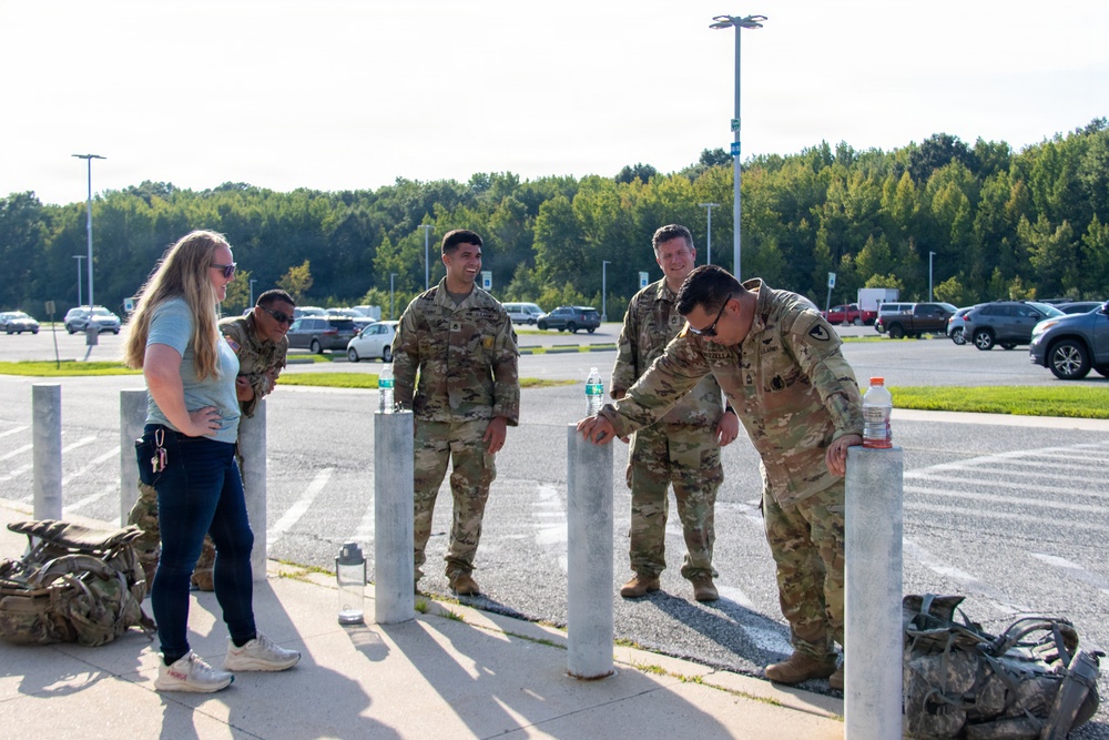 A group of five individuals, four in U.S. Army camouflage uniforms (OCP pattern) and one civilian woman in casual clothing, are gathered near concrete bollards in a parking lot area. They appear to be in a relaxed, social interaction, possibly during a break from physical training or a ruck march event, as military rucksacks are visible on the ground. The setting appears to be a military installation or government facility parking lot.