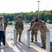 A group of five individuals, four in U.S. Army camouflage uniforms (OCP pattern) and one civilian woman in casual clothing, are gathered near concrete bollards in a parking lot area. They appear to be in a relaxed, social interaction, possibly during a break from physical training or a ruck march event, as military rucksacks are visible on the ground. The setting appears to be a military installation or government facility parking lot.