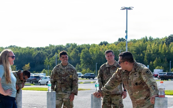 A group of U.S. Army soldiers in OCP (Operational Camouflage Pattern) uniforms are gathered near bollards in what appears to be a military installation parking lot. They are interacting with a civilian woman in casual clothing, appearing relaxed and engaged in conversation. Military rucksacks are visible on the ground nearby, suggesting a recent or upcoming ruck march or physical training event.