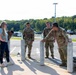 A group of U.S. Army soldiers in OCP (Operational Camouflage Pattern) uniforms are gathered near bollards in what appears to be a military installation parking lot. They are interacting with a civilian woman in casual clothing, appearing relaxed and engaged in conversation. Military rucksacks are visible on the ground nearby, suggesting a recent or upcoming ruck march or physical training event.
