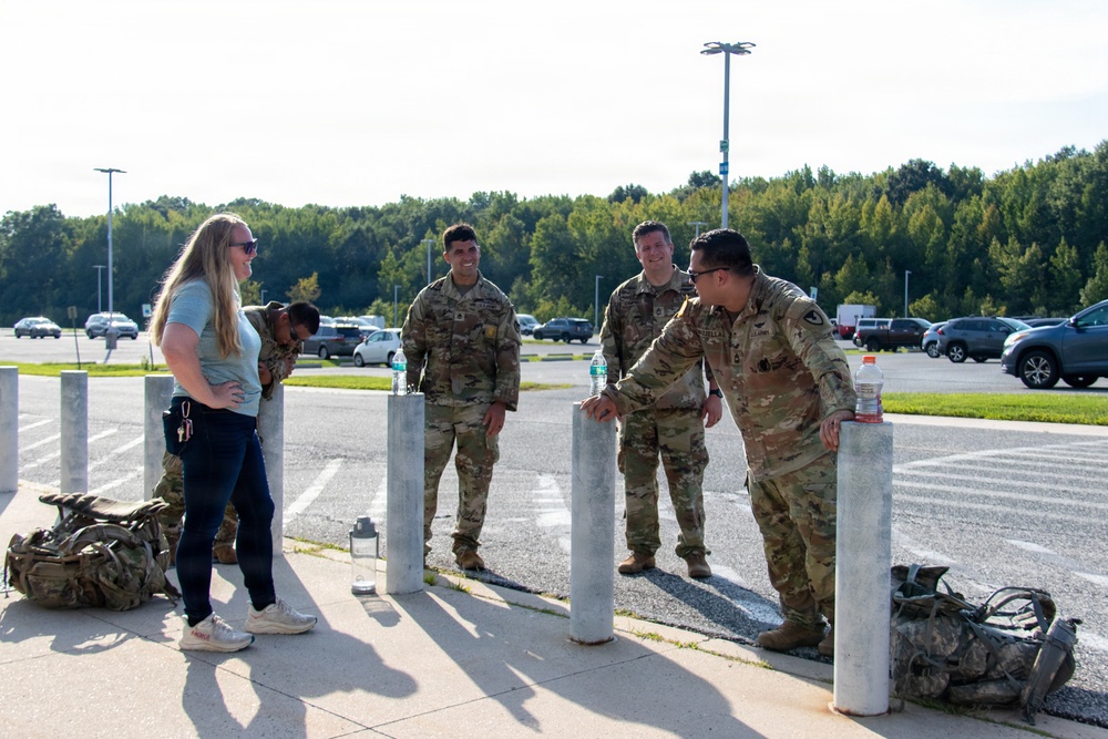A group of military personnel in OCP (Operational Camouflage Pattern) uniforms are gathered near concrete bollards in what appears to be a military installation parking lot. They are interacting with a civilian woman in casual clothing. Military rucksacks are visible on the ground nearby, suggesting a ruck march or physical training event.