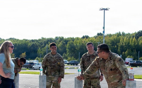 A group of military personnel in OCP (Operational Camouflage Pattern) uniforms are gathered near concrete bollards in what appears to be a military installation parking lot. They are interacting with a civilian woman in casual clothing. Military rucksacks are visible on the ground nearby, suggesting a ruck march or physical training event.