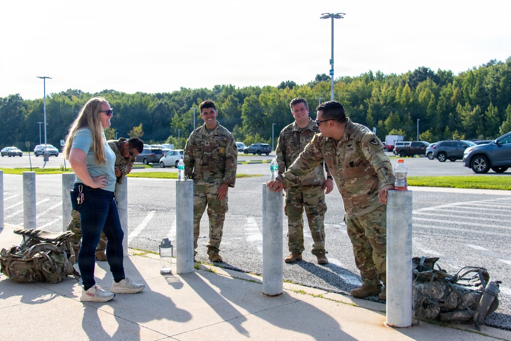 A group of U.S. Army soldiers in OCP (Operational Camouflage Pattern) uniforms are gathered near bollards in a parking lot, conversing with a civilian woman in casual clothing. Military rucksacks are visible on the ground nearby, suggesting the group may have recently completed or is preparing for a ruck march or physical training event. The setting appears to be a military installation or base facility during daytime.