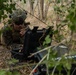 A U.S. Marine in full combat gear is lying prone in a wooded, brushy area while operating what appears to be military communications or electronic equipment housed in a ruggedized case. The soldier is wearing MARPAT camouflage, a tactical helmet with night-vision mount, and sunglasses, suggesting a field training exercise or operational deployment. The setting appears to be a dry, sparse woodland environment.