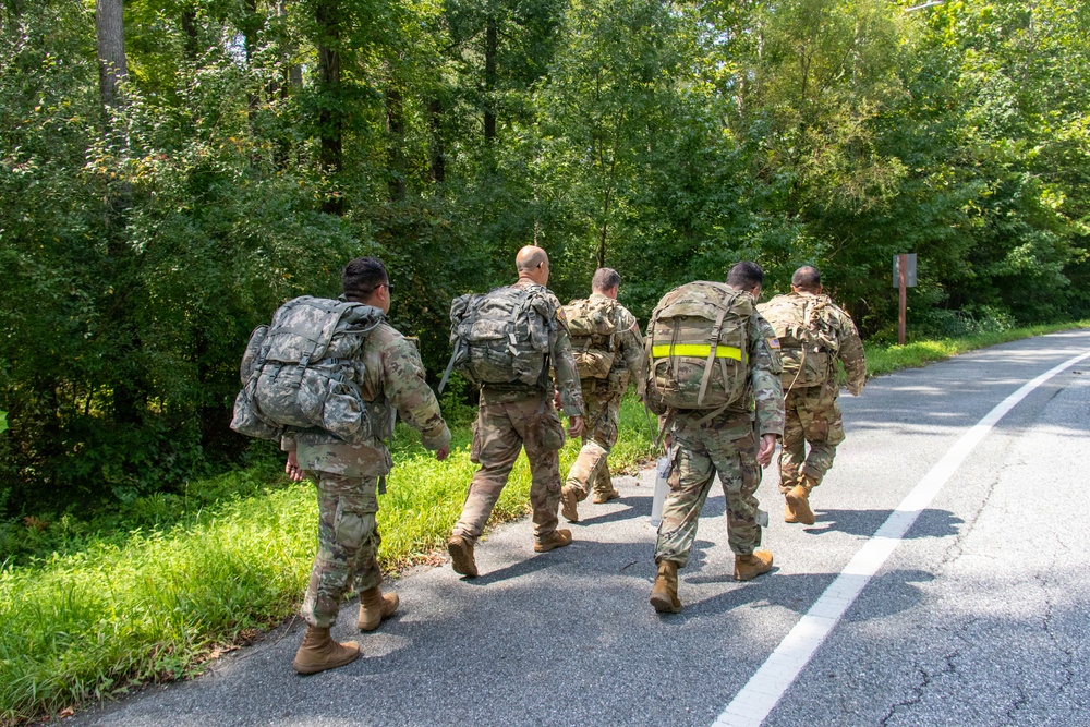 A group of five military personnel in camouflage uniforms are conducting a road march along a paved road bordered by dense green vegetation. They are all carrying large rucksacks and wearing standard-issue combat boots, moving away from the camera. One soldier has a high-visibility yellow reflective band on their pack, likely for safety identification.