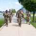 A group of five military personnel in U.S. Army OCP (Operational Camouflage Pattern) uniforms are walking along a sidewalk on what appears to be a military installation. They are carrying rucksacks and tactical gear, suggesting they are conducting or completing a ruck march or physical training exercise. The setting includes well-maintained grounds, modern buildings, and a flagpole in the background.