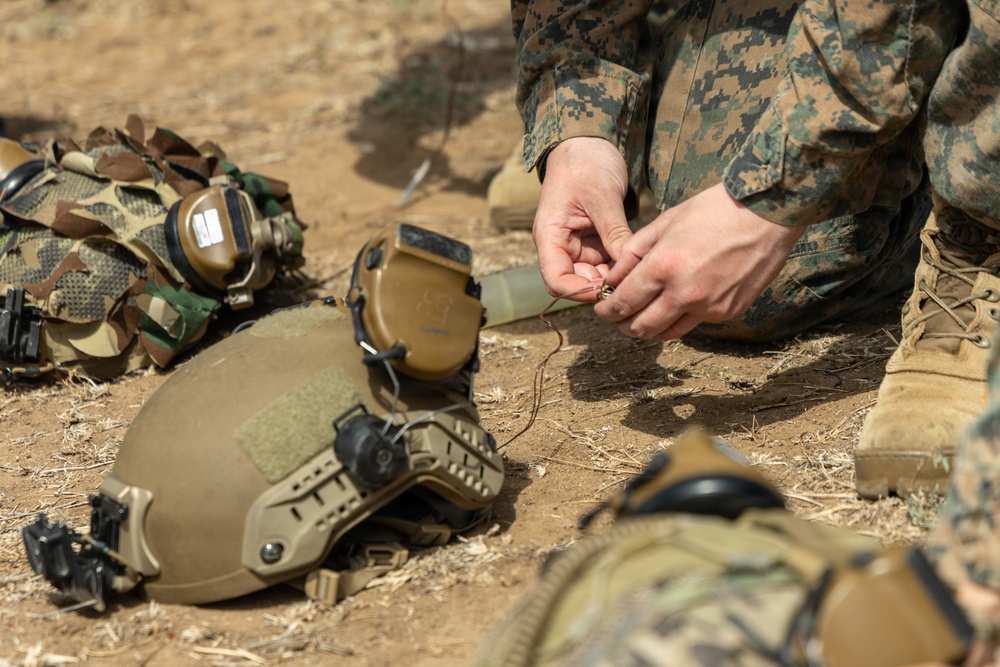 Military personnel in digital camouflage uniforms are gathered on arid ground, handling small components or wire with their hands. Multiple tactical helmets with communication headsets and rail-mounted accessories are visible on the ground. The scene appears to depict a training or preparation activity in a field environment.